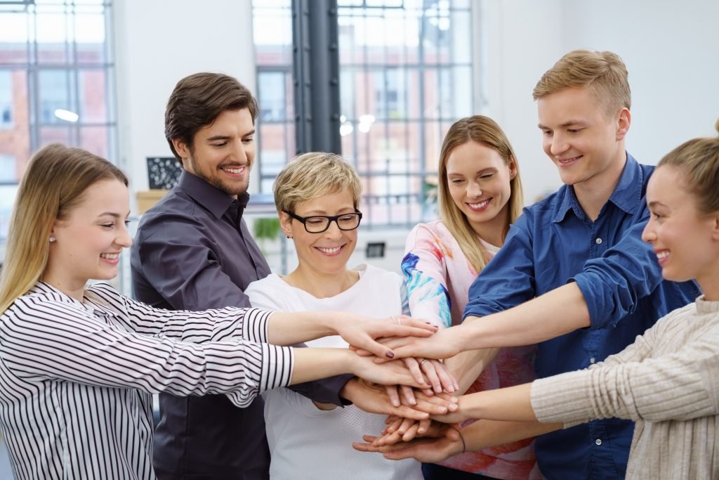 Six happy, smiling work colleagues participating in a hands in team-building exercise in an office room.